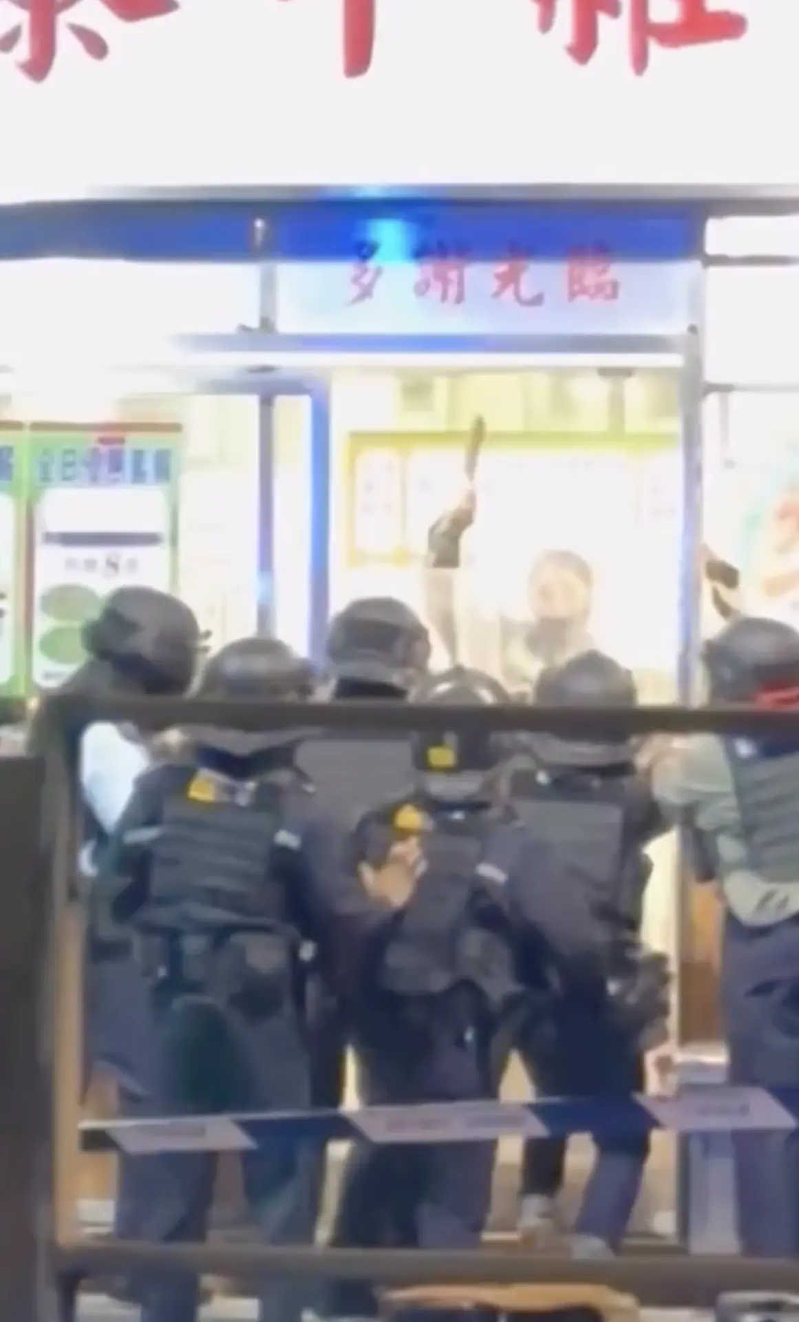 網上流傳多段現場片段可見，涉事女子身穿淺色牛仔褸、佩戴黑色口罩，先後持剪刀及菜刀隔著店舖窗口，與大批戒備警員對峙。畫面顯示，至少 6 名身穿戰術背心、佩戴頭盔的軍裝警員，手持警棍及透明長盾在店門戒備。(圖：視頻截圖)